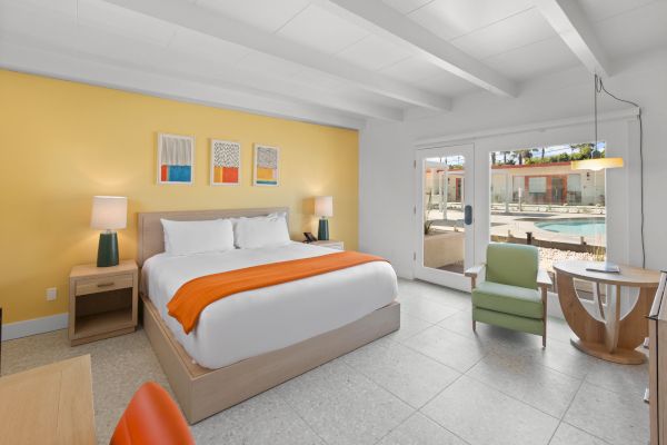 A modern hotel room features a king-sized bed with an orange throw, two bedside tables with lamps, a small seating area, and a pool view through glass doors.