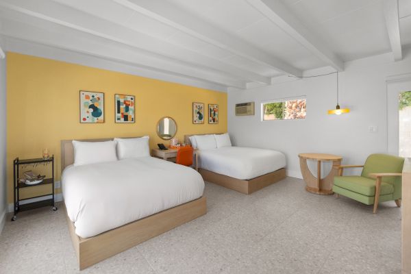 A modern, minimalist room with two beds, yellow accent wall, wall art, a desk with a mirror, a green chair, and a small round table.