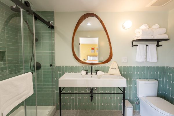 Modern bathroom with a shower, sink, mirror, and toilet. Green tiles, towels on a shelf, and wall light complete the design.