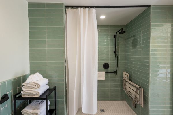 A bathroom with green tiled walls, a shower curtain, a black showerhead, a black metal towel shelf with white towels, and a foldable shower seat.