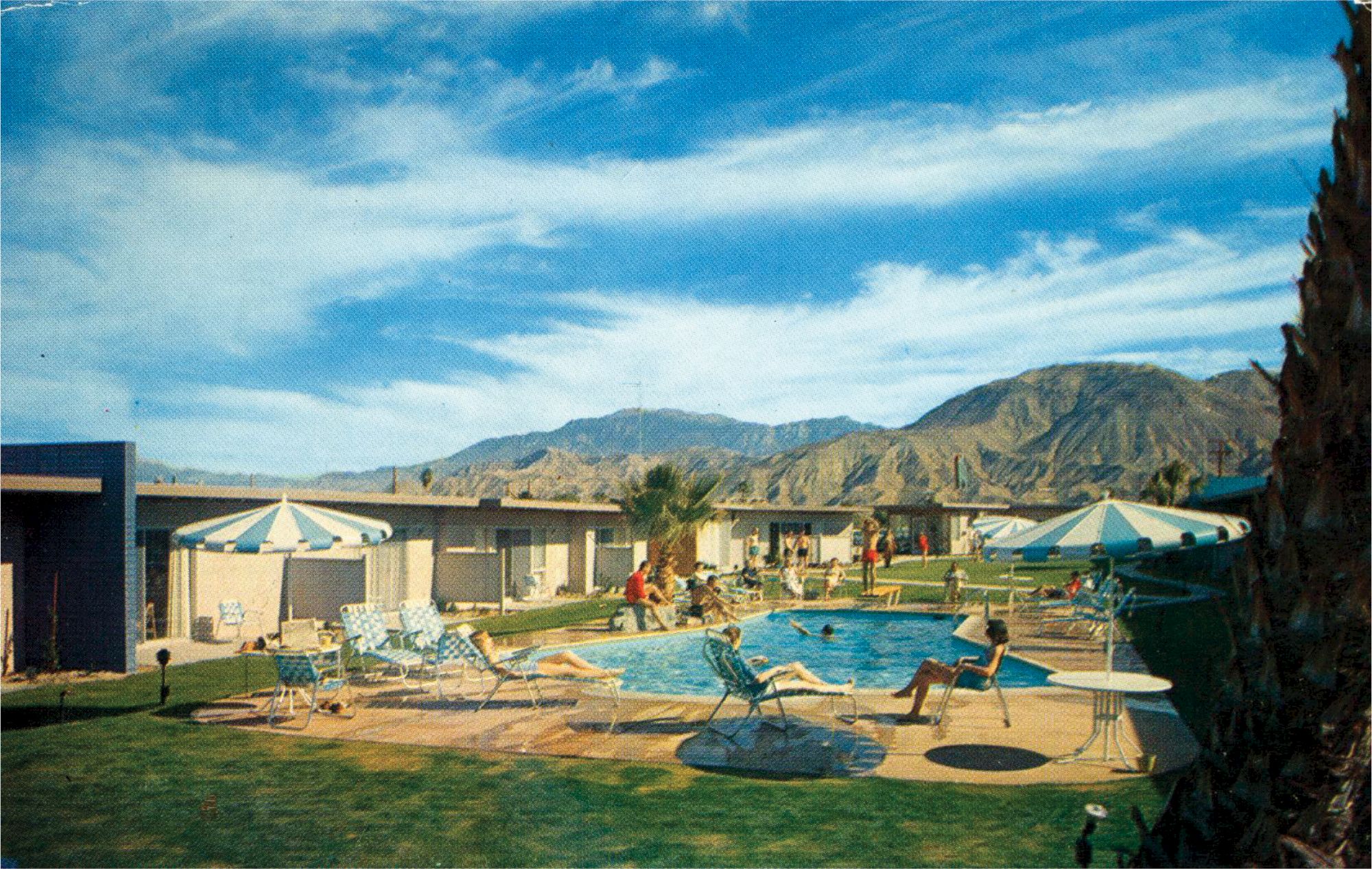 A swimming pool area with people relaxing, surrounded by umbrellas and chairs, with mountains in the background and a clear sky above.