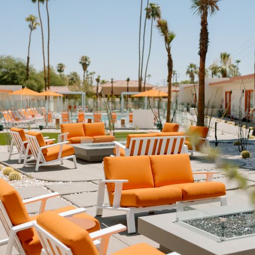 Outdoor seating area with orange couches and white-framed chairs, fire pits, and tall palm trees under a clear blue sky.