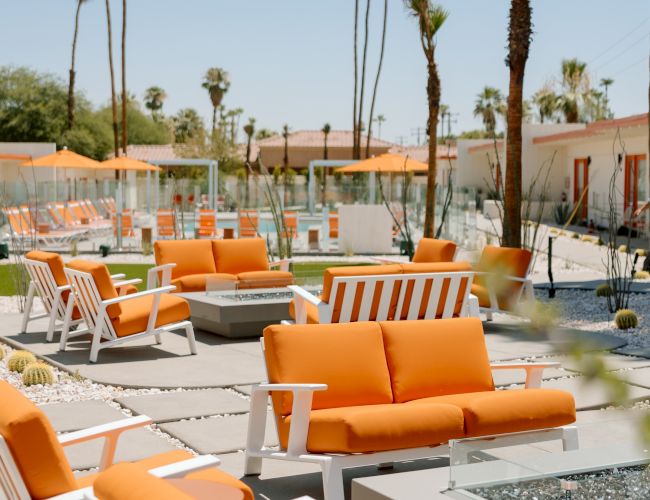 Outdoor seating area with orange couches and white-framed chairs, fire pits, and tall palm trees under a clear blue sky.