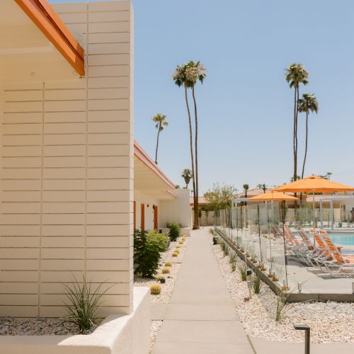 A modern walkway between buildings with a poolside area, orange parasol-covered chairs, palm trees, and a clear sky in the background.