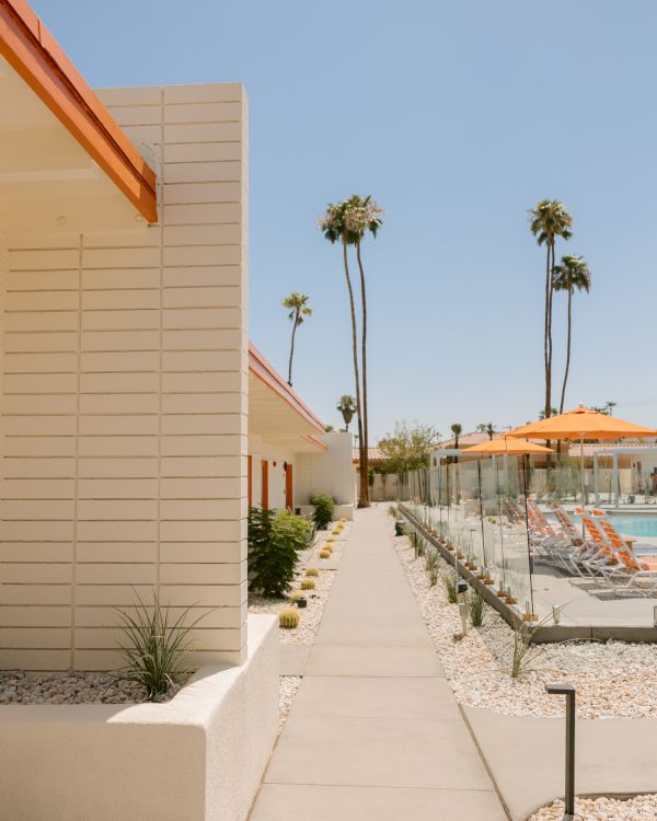 A modern walkway between buildings with a poolside area, orange parasol-covered chairs, palm trees, and a clear sky in the background.