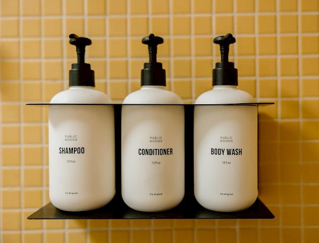 The image shows three white pump bottles labeled as shampoo, conditioner, and body wash placed on a shelf, in front of a tiled wall.