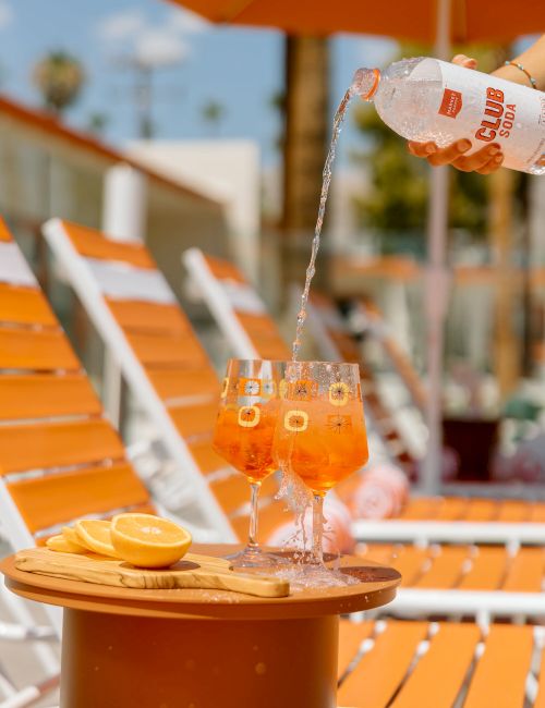 The image shows glasses of drinks being poured, with orange slices on a table by orange lounge chairs and umbrellas at a poolside setting.