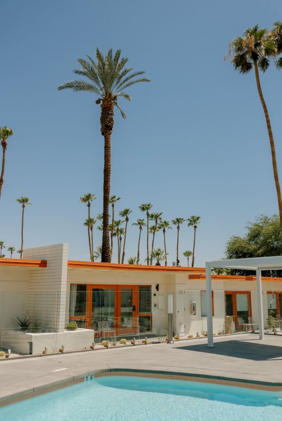 This image shows a modern building with large windows, surrounded by tall palm trees and a swimming pool, under a clear blue sky.