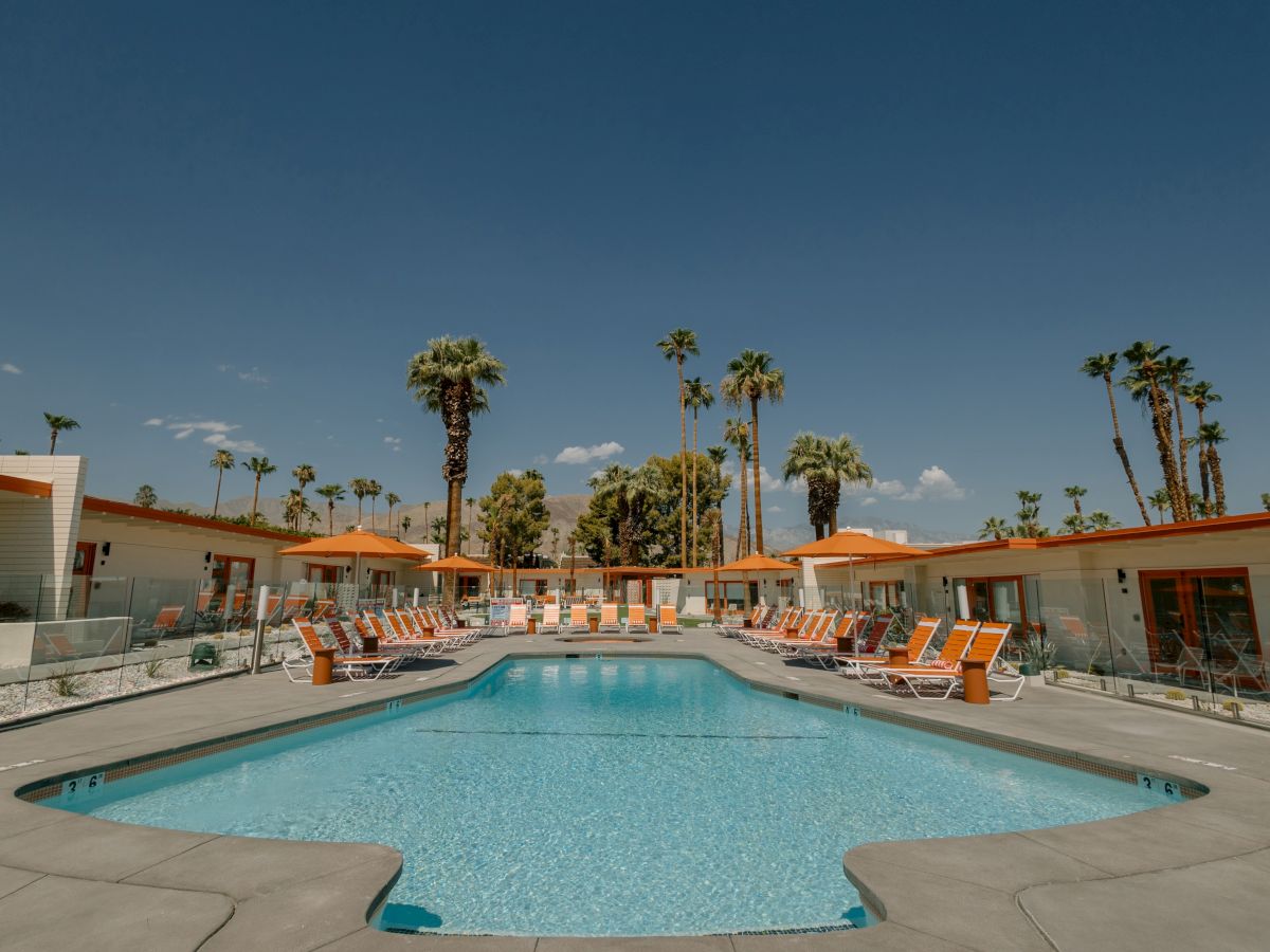 A swimming pool surrounded by lounge chairs is set in the center of a mid-century modern style hotel courtyard with palm trees and a clear sky.