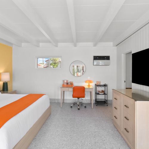 The image shows a modern bedroom with a king-sized bed, orange accents, a desk, chair, lamp, mirror, dresser, and a wall-mounted TV.