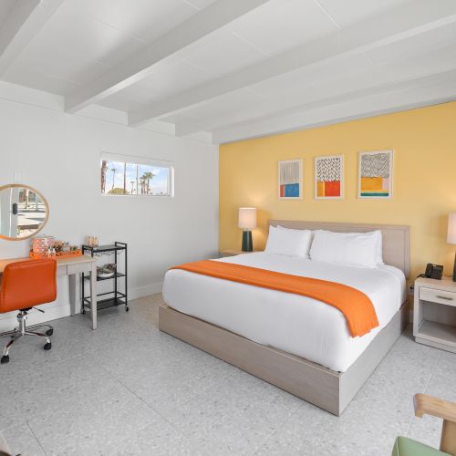 A modern bedroom with a double bed, orange accents, desk with chair, table lamps, wall art, and a window. The room has a bright, cozy feel.