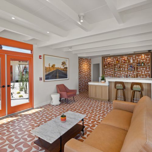 A modern living space features an orange accent theme, a sofa, two armchairs, a small bar area, and geometric patterned tiles under a white ceiling.