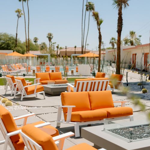 The image shows an outdoor seating area with orange cushioned chairs and sofas, fire pits, and palm trees on a sunny day, creating a relaxed ambiance.