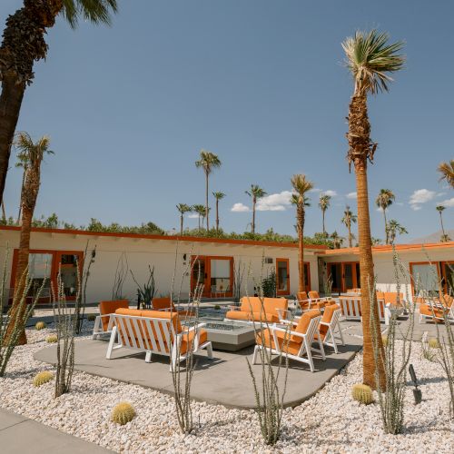 The image shows an outdoor patio area with orange and white furniture, surrounded by palm trees and desert plants under a clear sky.