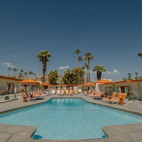 This image features a swimming pool surrounded by chairs and tables with umbrellas. Palm trees and a clear sky are visible in the background.