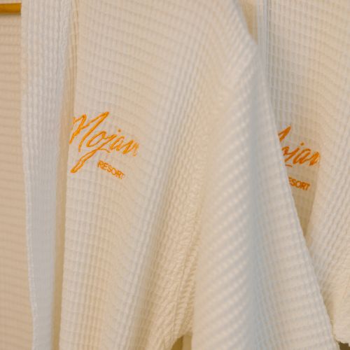 The image shows two white waffle-knit bathrobes hanging, embroidered with the name 