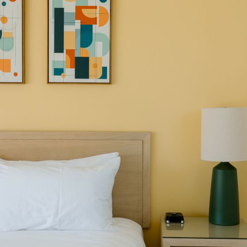 The image shows a bedroom with a bed, white pillow, nightstand, modern table lamp, and two abstract geometric art pieces on a yellow wall.