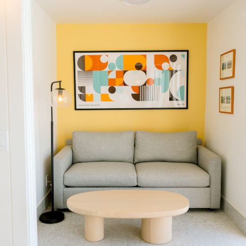 A cozy room with a gray sofa, colorful abstract wall art, yellow accent wall, floor lamp, and small wooden coffee table.