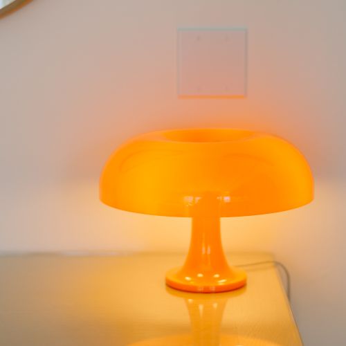 An orange table lamp is placed on a glass surface, its reflection visible below. The light creates a warm glow on the nearby wall.