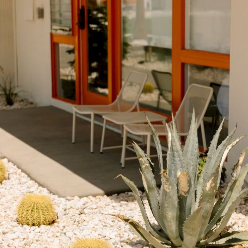 Outdoor patio with two metal chairs, orange-framed doors, and arid landscaping featuring cacti and succulents among white pebbles.