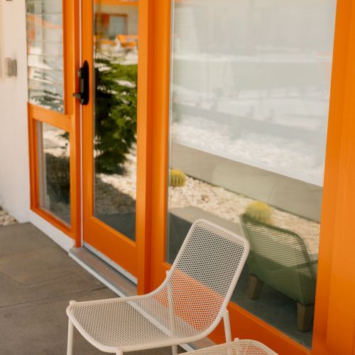 The image shows a bright orange-framed glass door with a white curtain, and two white mesh chairs in front of it on a gray concrete floor.