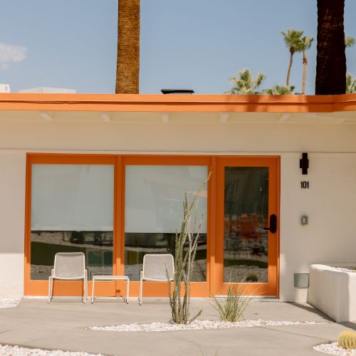 The image shows a modern house with an orange trim, large glass doors, two chairs on the patio, white gravel, and green lawn.