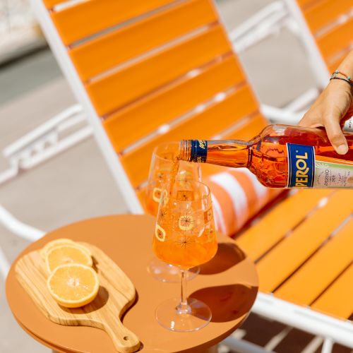 A bottle pouring Aperol into a glass on a table next to a cutting board with orange slices; with two orange lounge chairs in the background.