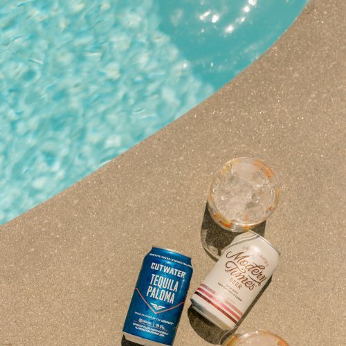 Cans of beverages and two drinks rest on the edge of a pool beside clear, blue water, casting reflections in the sun.