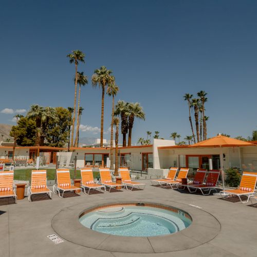 An outdoor spa area with orange lounge chairs and umbrellas, surrounded by palm trees and buildings under a clear blue sky.