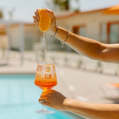 A person squeezes an orange into a cocktail glass near a pool, suggesting a refreshing summer drink by the poolside on a sunny day.