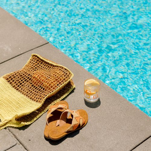 A yellow woven bag, brown sandals, and a jar of cream are placed on a poolside pavement beside a clear blue swimming pool.