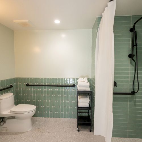 The image shows a modern bathroom with green tiling, a toilet, a curtained shower, and a black towel rack holding white towels.