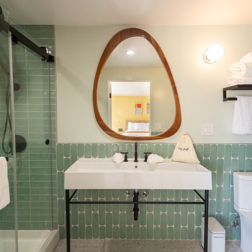 A modern bathroom with a shower, sink, toilet, and mirror. Tiles are green, towels are neatly stacked, and the design is clean and minimalist.