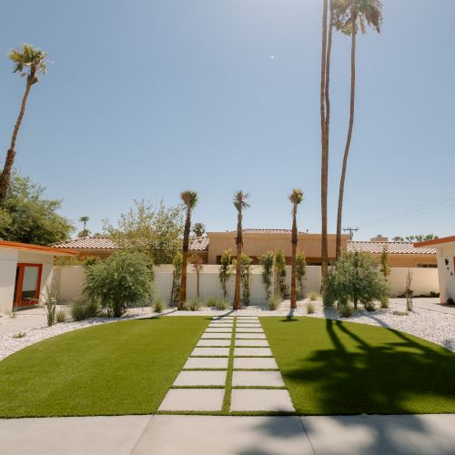 A landscaped courtyard with artificial grass, paver path, palm trees, and desert plants, surrounded by white walls with red-framed doors.